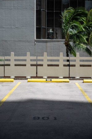 A parking area with a white fence, yellow parking lines, and a building with large windows in the background. - Just Renovated Marine Surf Corner Unit with Free Parking! (Honolulu)