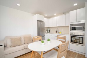 A modern kitchen with white cabinets, a dining table with chairs, a sofa, and a built-in oven. - Just Renovated Marine Surf Corner Unit with Free Parking! (Honolulu)