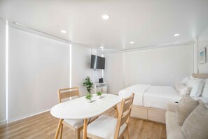 A modern, minimalist room with a white table and chairs, a bed with a white duvet, and a flat-screen TV mounted on the wall. - Just Renovated Marine Surf Corner Unit with Free Parking! (Honolulu)