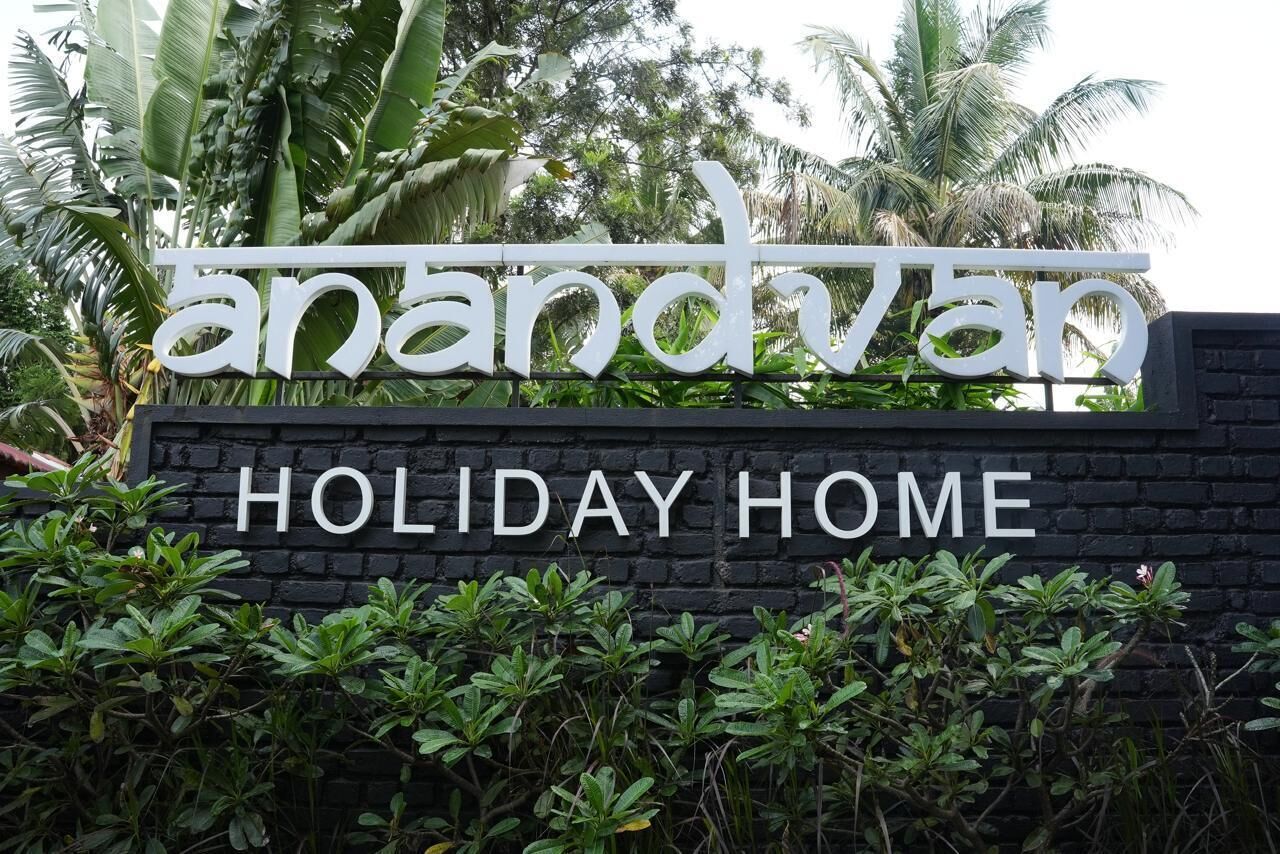 A sign for 'Anandvan Holiday Home' with a black brick wall and lush greenery in the background.