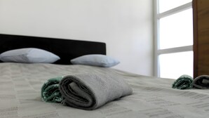 A neatly made bed with a grey blanket and rolled-up green towel, a black headboard, and a window with daylight.