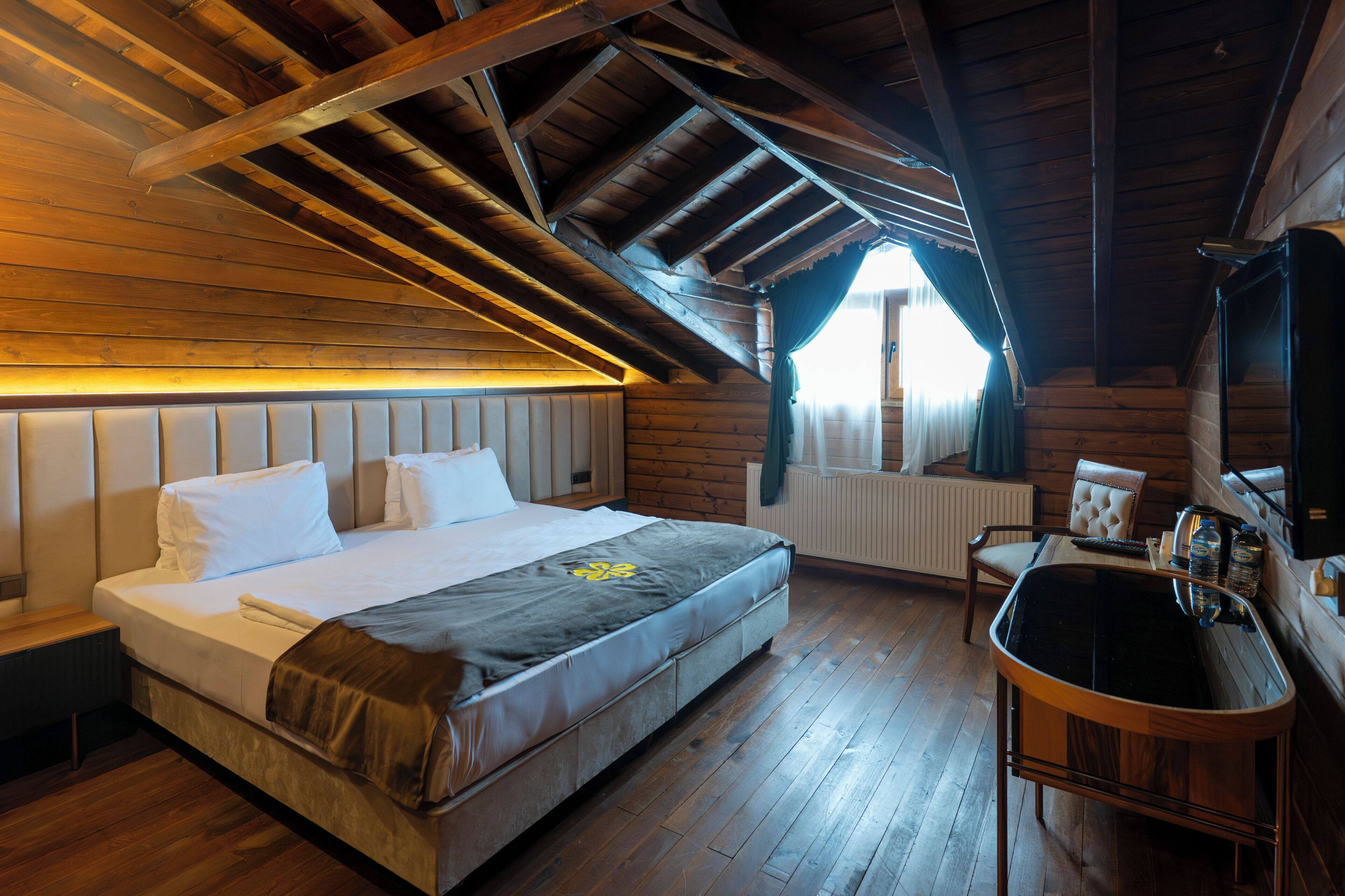 Um quarto com uma cama grande, teto de madeira, janela com cortinas, radiador e uma pequena mesa com chaleira e garrafas.