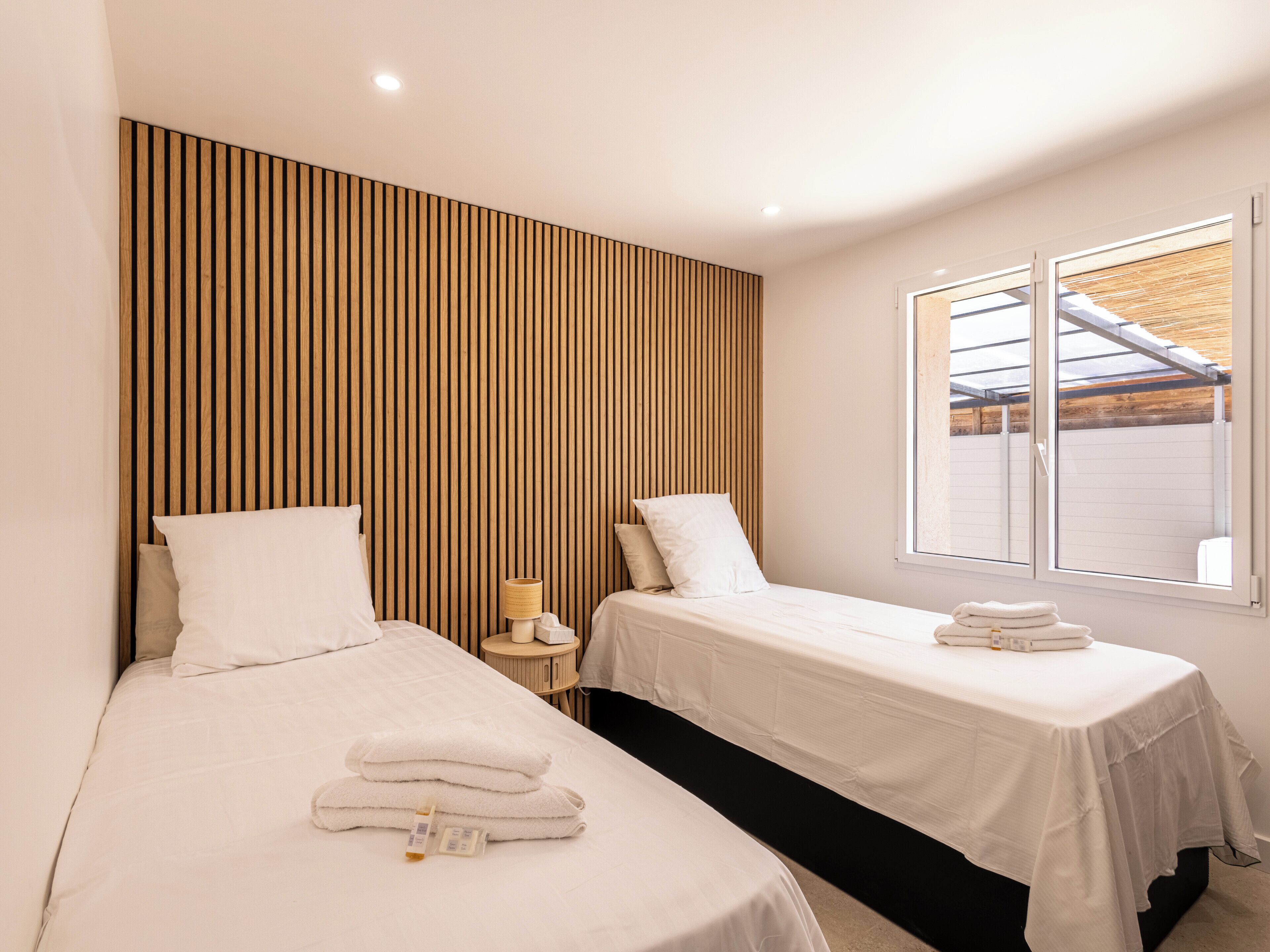 A bedroom with two beds, a wooden slat headboard, a bedside table with a lamp, and a window with a view of the outside.