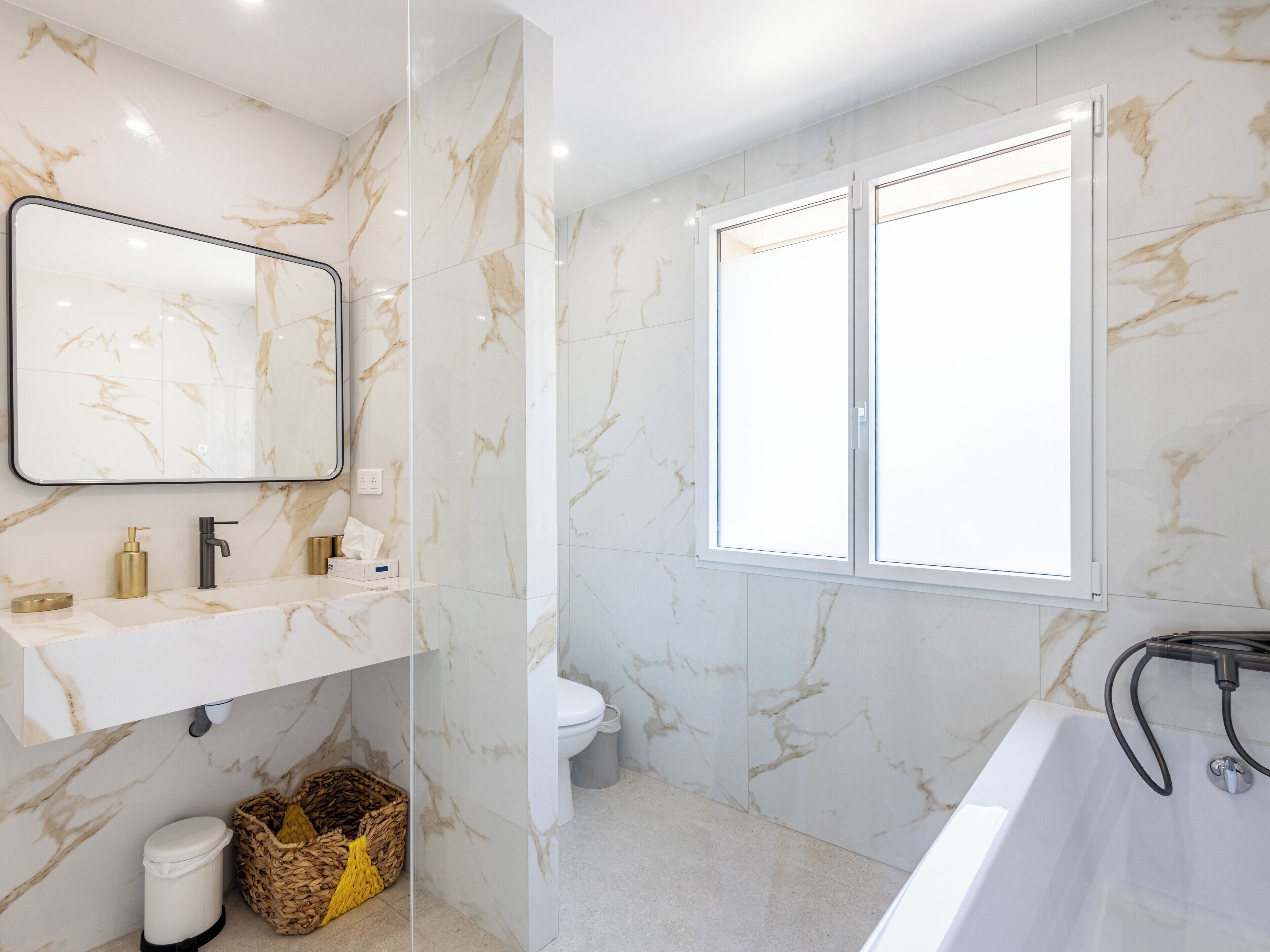 Een badkamer met een marmeren wand, een toilet, een wastafel met spiegel en een bad.