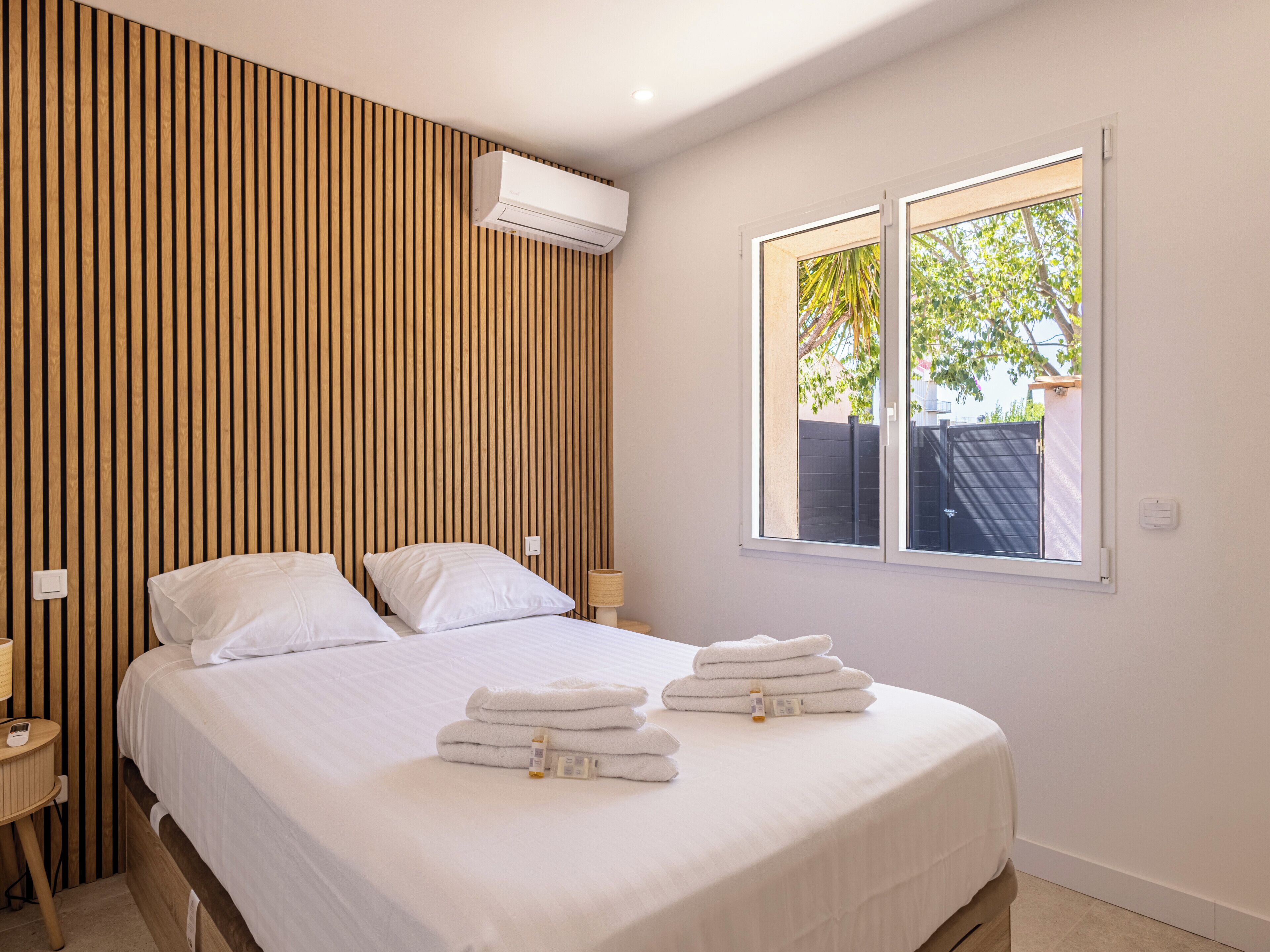 A hotel room with a bed, a window, a wall-mounted air conditioner, and a bedside table with a lamp.