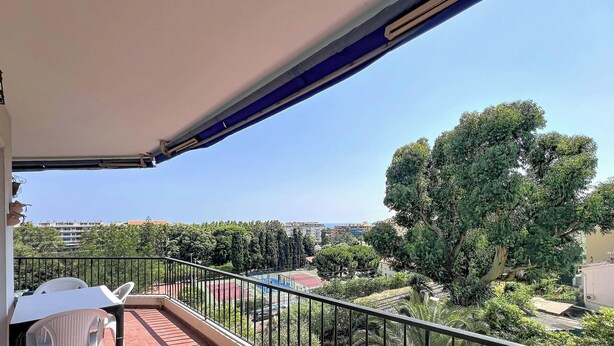 A balcony with a white umbrella, a white chair, and a metal railing. In the background, there are trees and a sports court. - Spacious studio, terrace, sea view - 2 guests (Menton)