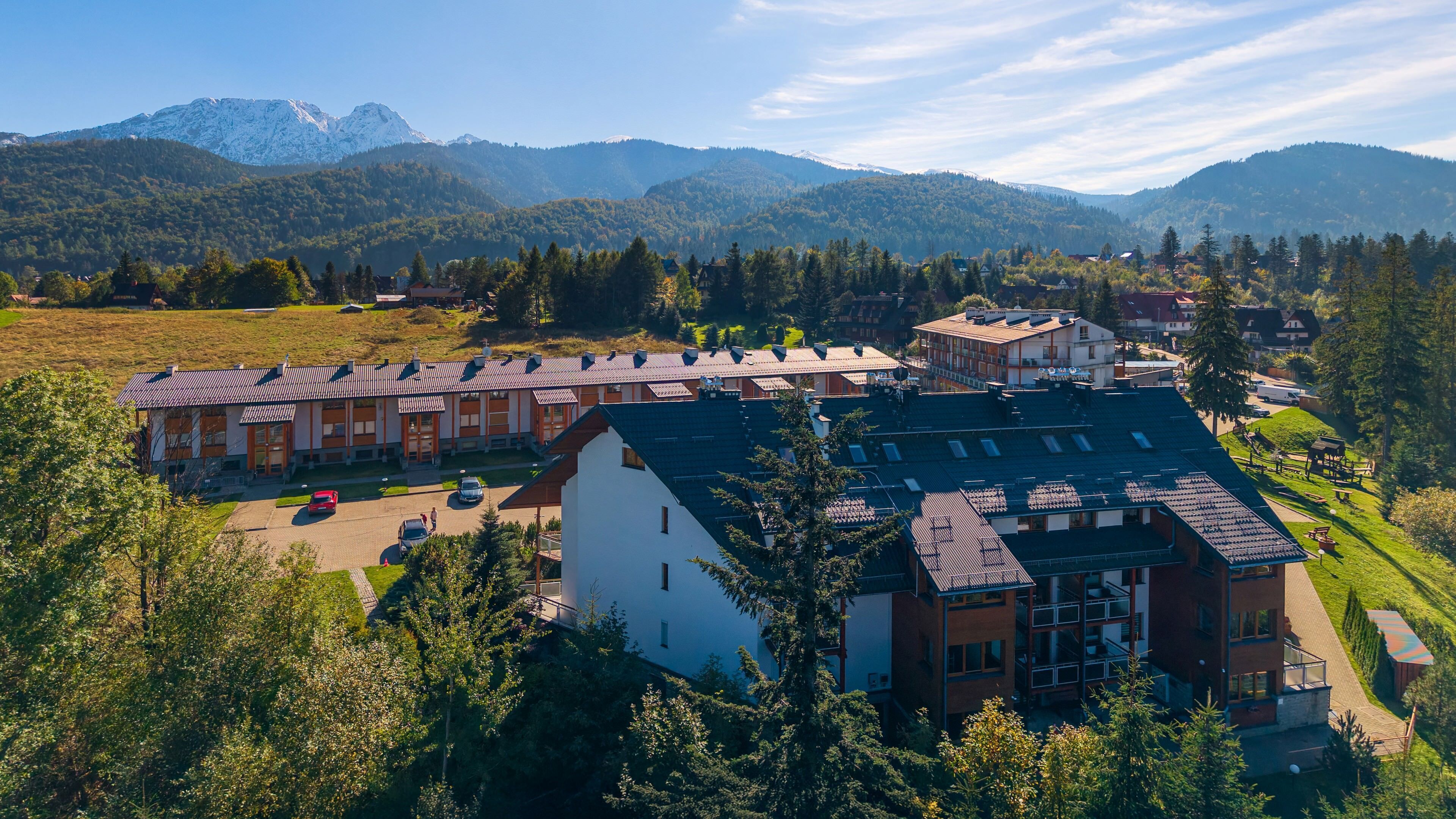 A modern hotel with multiple buildings, a parking area, and a mountainous backdrop.