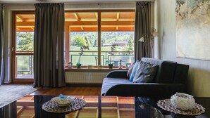 A room with a sofa, a table, and a view of a garden through large windows. - Apartamenty Sun&Snow VIVA MARIA SPA II (Zakopane)