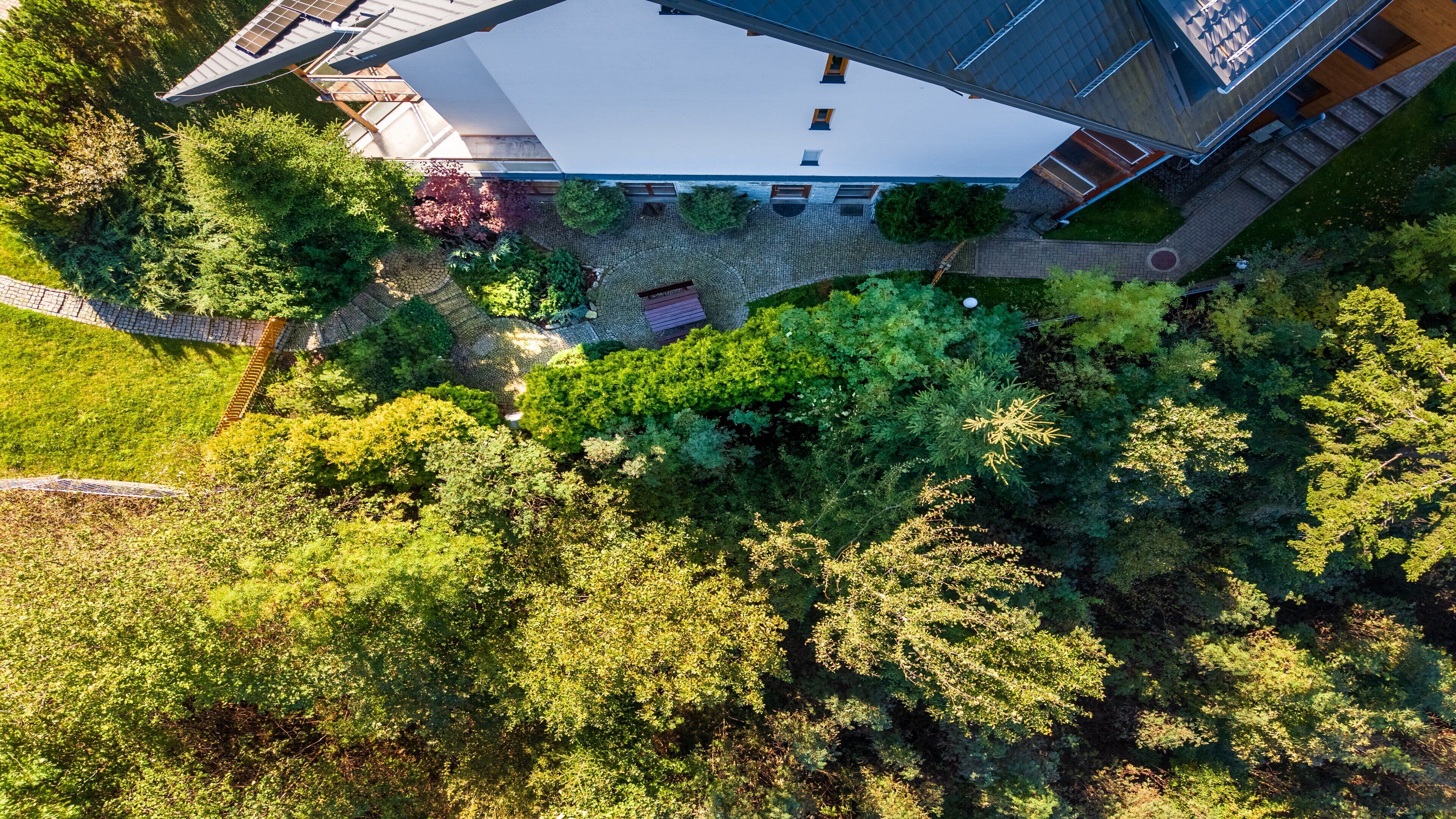 An aerial view of a modern house with a large white roof, surrounded by lush greenery and a stone pathway.