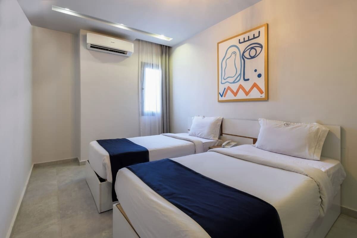Une chambre d’hôtel avec deux lits, une fenêtre avec des rideaux, un appareil de climatisation et une œuvre d’art accrochée au mur.
