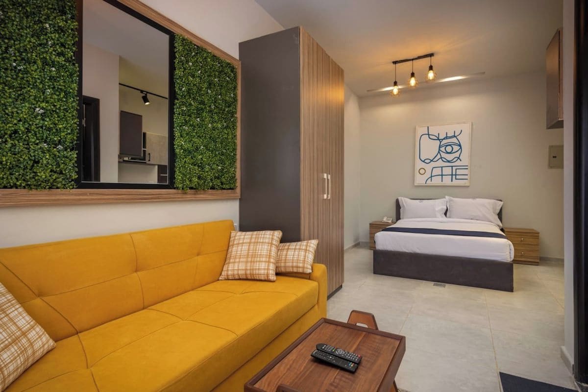 A modern hotel room with a yellow sofa, a bed, a mirror, a green wall, and a painting.