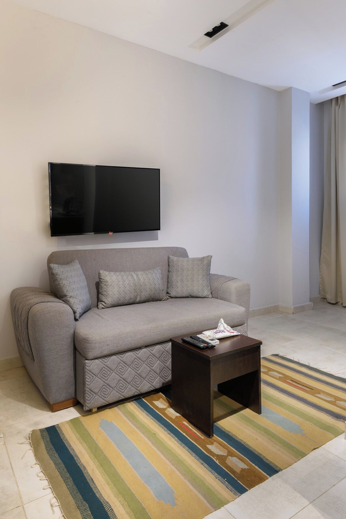 A living room with a sofa, a coffee table, a flat-screen TV mounted on the wall, and a striped rug on the floor.