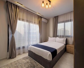 A modern hotel room with a large bed, a bedside table, an air conditioning unit, and a view of the city through sheer curtains. - Golden Gates Residence (El Mokkatam)