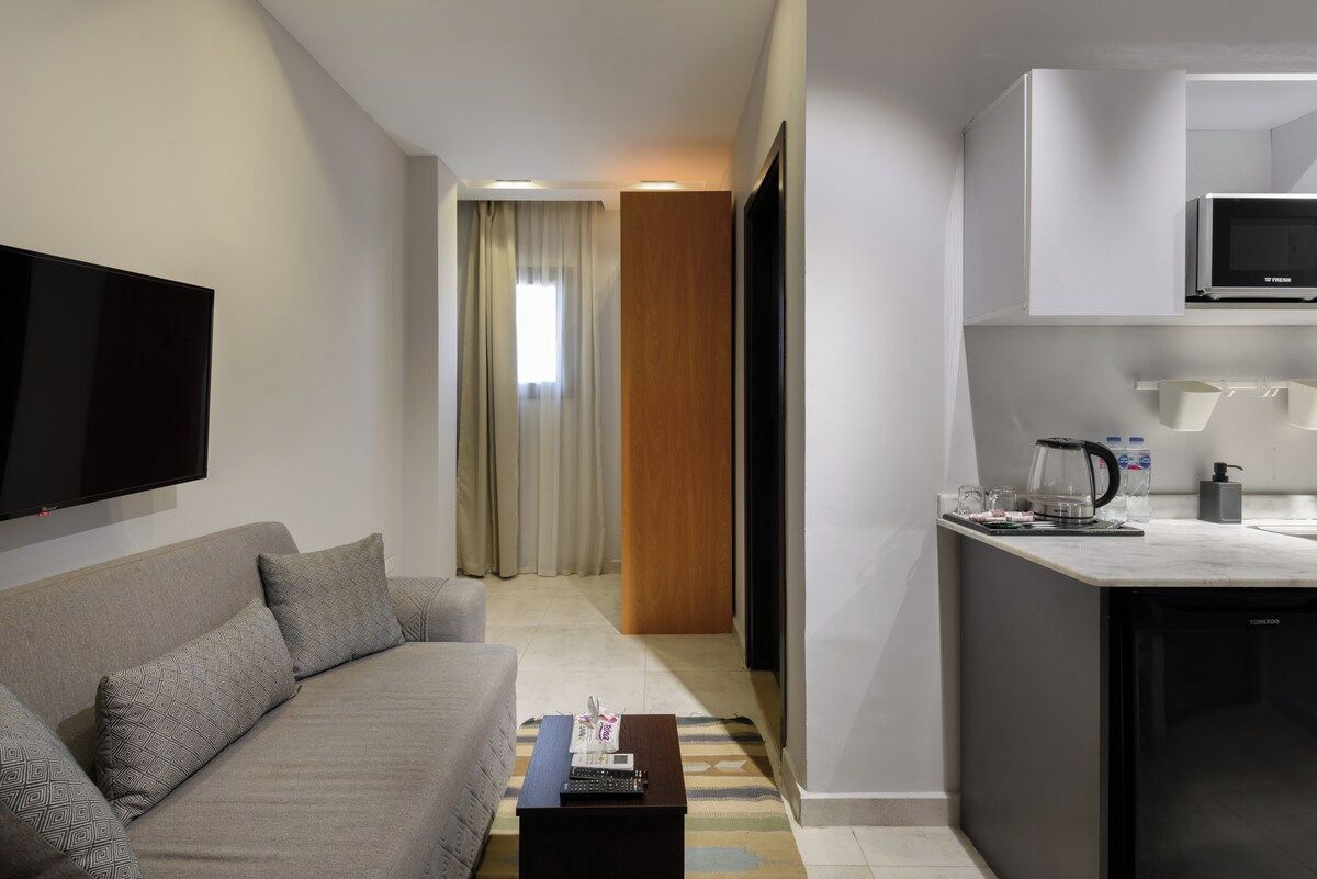 A modern hotel room with a sofa, a coffee table, a flat-screen TV, a kitchenette with a microwave, and a sliding door leading to a bathroom.