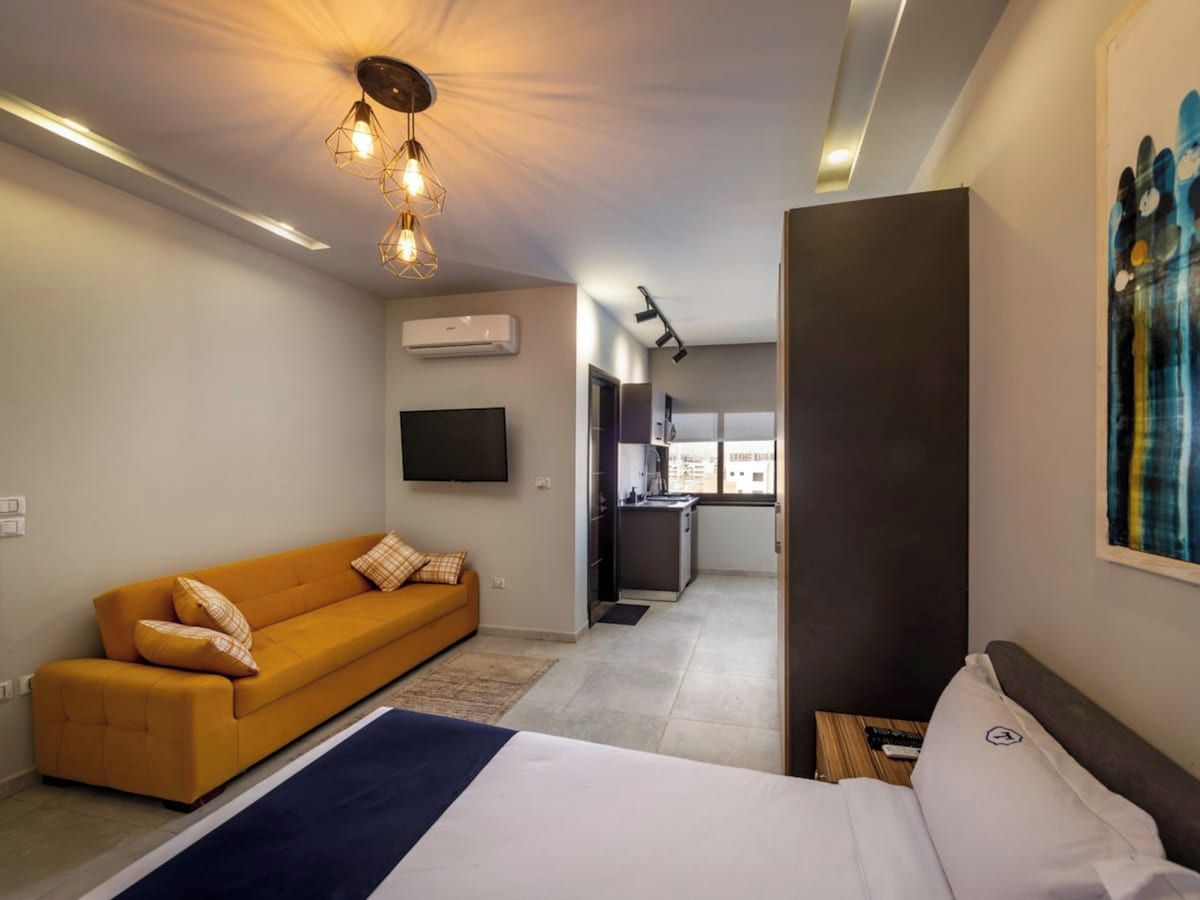 Une chambre d’hôtel moderne avec un canapé, un lit, un téléviseur à écran plat et un coin cuisine à l’arrière-plan.