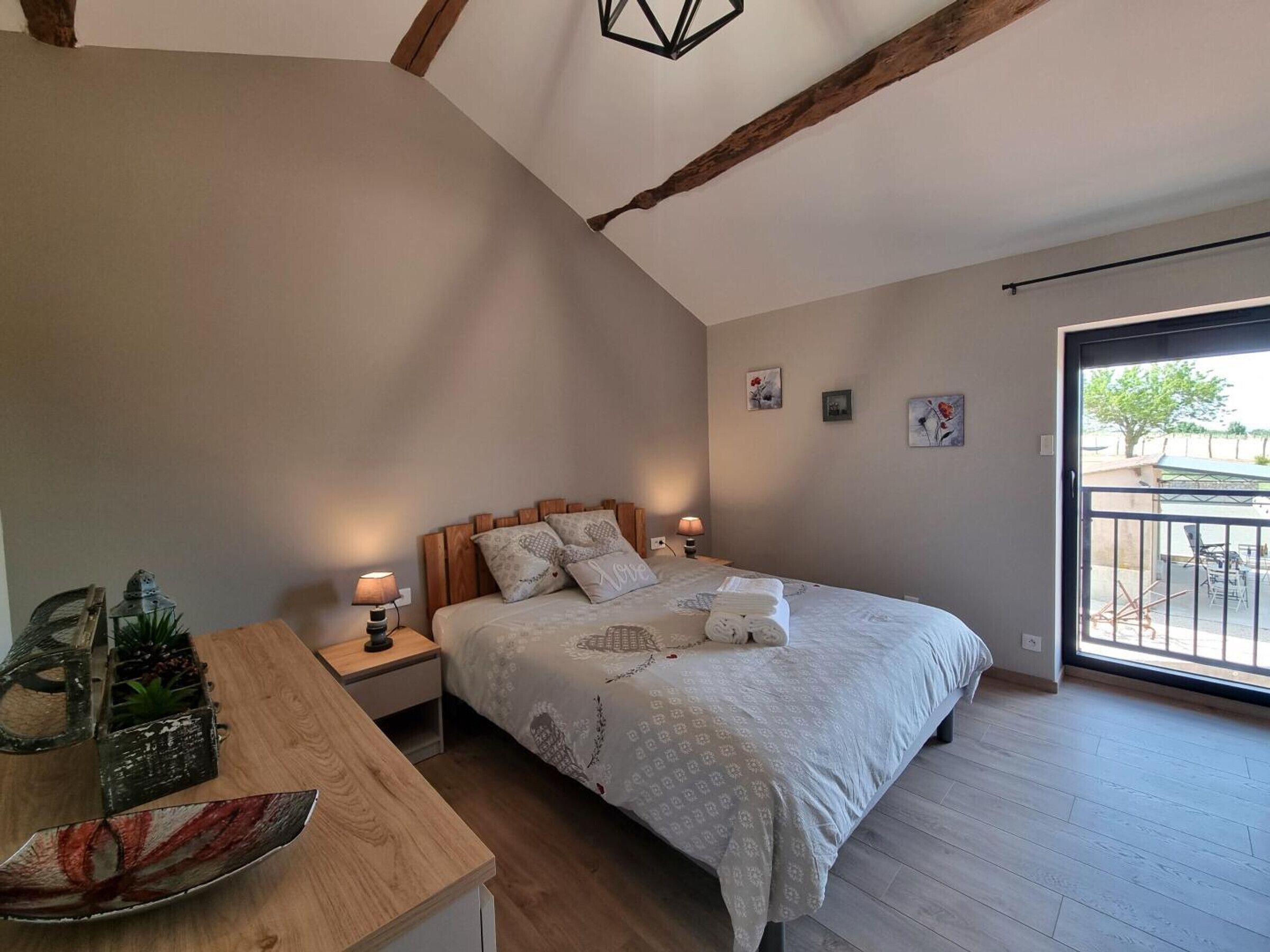 A bedroom with a large bed, a wooden nightstand, a desk with a plant, and a view of the outside through a glass door.
