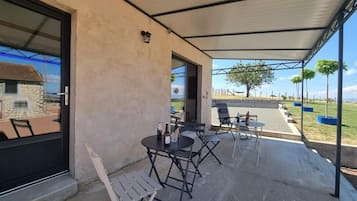 Un área de estar al aire libre con techo, mesas y sillas, vista a un paisaje rural y un edificio de piedra al fondo.