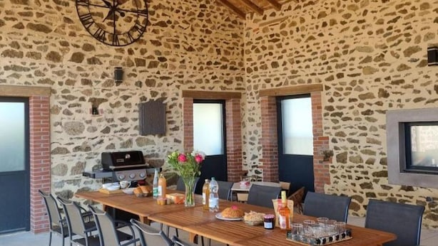 Un espacio al aire libre para comer con paredes de piedra, una mesa de madera, sillas, una parrilla y un reloj.