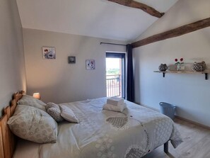 Quarto com cama, almofadas, mesa de cabeceira com luminária, janela com vista e prateleira com objetos decorativos.