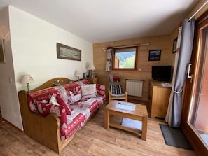 Ein gemütliches Zimmer mit einer Holzcouch, einem kleinen Tisch, einem Heizkörper, einem Fernseher und einem Fenster mit Blick auf Grünflächen.