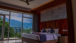 Une chambre avec un grand lit, un ventilateur de plafond, une vue sur les montagnes par la fenêtre et un balcon.