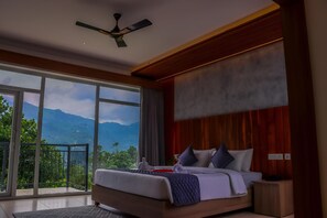 Une chambre avec un grand lit, un ventilateur de plafond, une vue sur les montagnes par la fenêtre et un balcon.