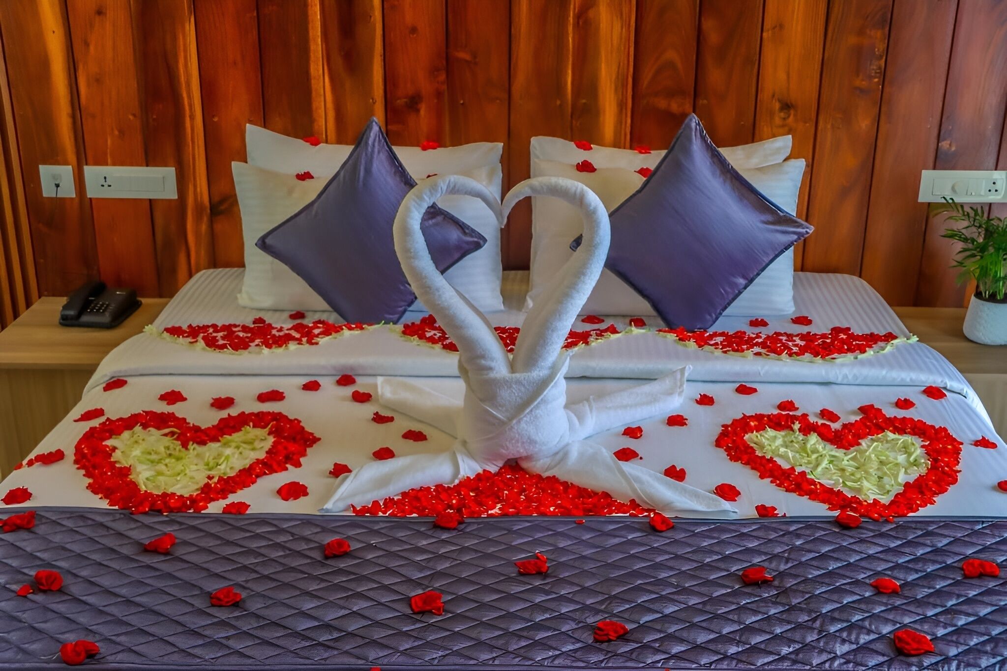 Una cama con una toalla dispuesta en forma de corazón, rodeada de pétalos de rosa roja, y dos almohadas con diseño de corazón.