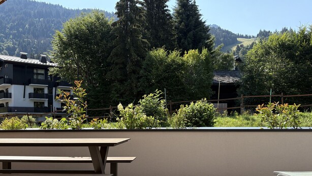 Eine Holzterrasse mit Picknicktisch und Liegestuhl mit Blick auf einen angelegten Garten und einen Berg im Hintergrund.