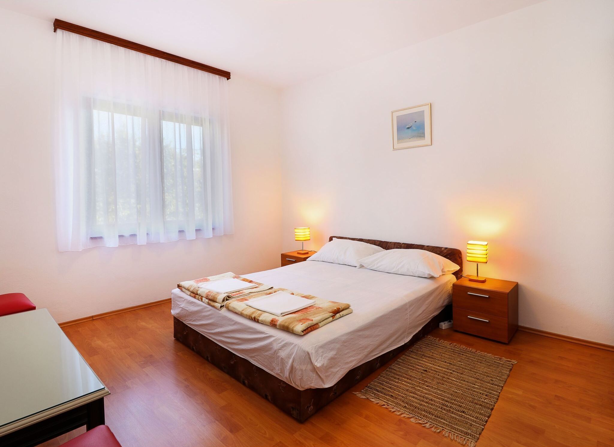 A neatly made bed with a floral bedspread, two bedside lamps, a small bedside table, a wooden nightstand, a framed picture on the wall, and a red chair.