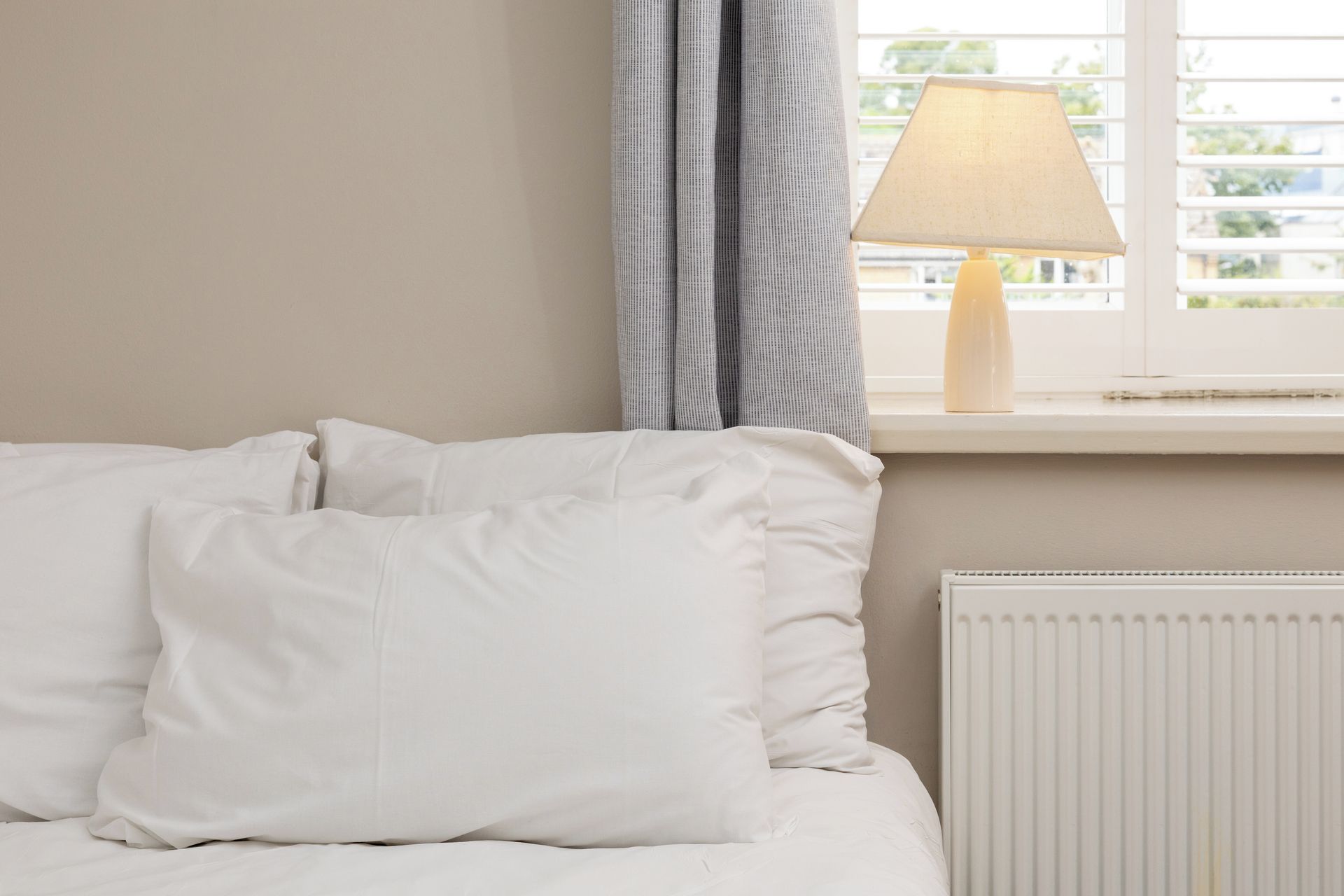 A bed with white bedding, a window with a view of greenery, and a lamp on the windowsill.