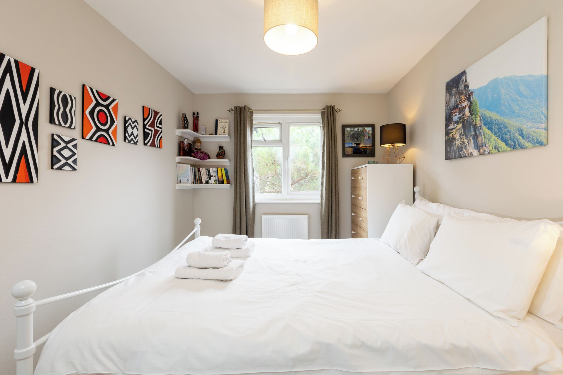 A bedroom with a bed, white bedding, a window with curtains, a shelf with books and decorative items, and a painting on the wall.