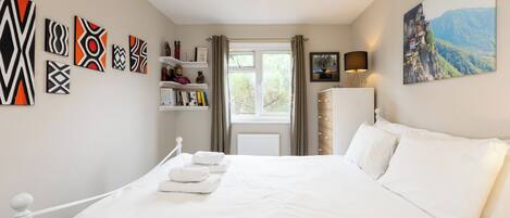 A bedroom with a bed, white bedding, a window with curtains, a shelf with books and decorative items, and a painting on the wall.
