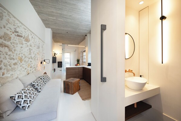 Un interior moderno con una pared de piedra, un sofá blanco con cojines estampados, una mesa de centro de madera y un baño con lavamanos y espejo blancos.