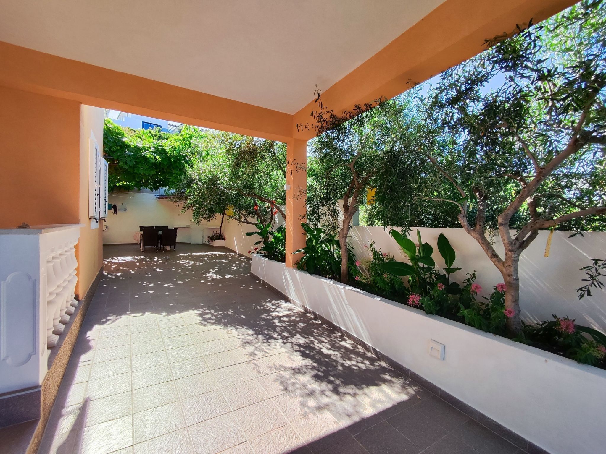 A covered outdoor area with a tiled floor, a seating arrangement, and a variety of plants and trees.