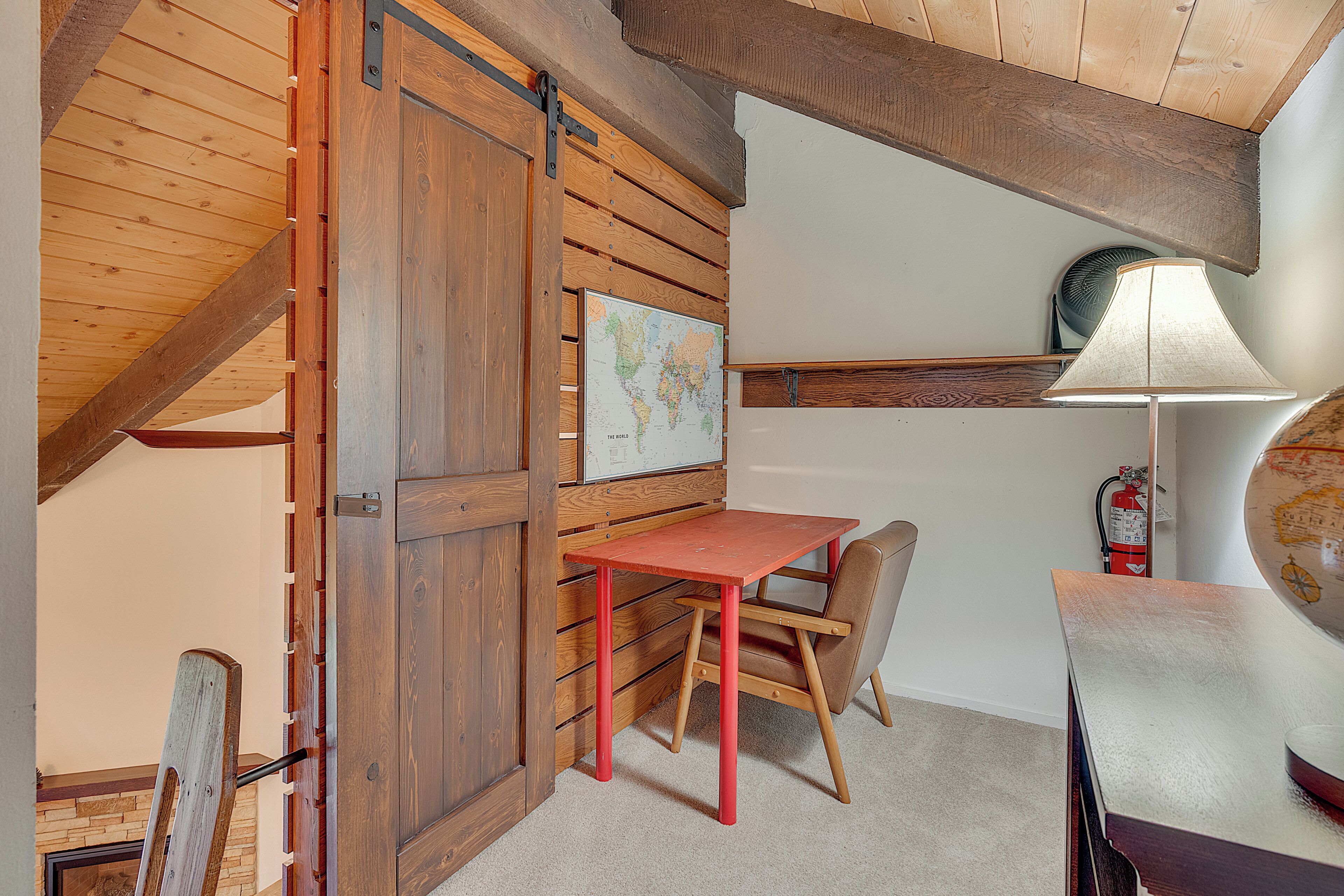 A room with a wooden sloped ceiling, a red table with a chair, a globe on the wall, and a lamp.
