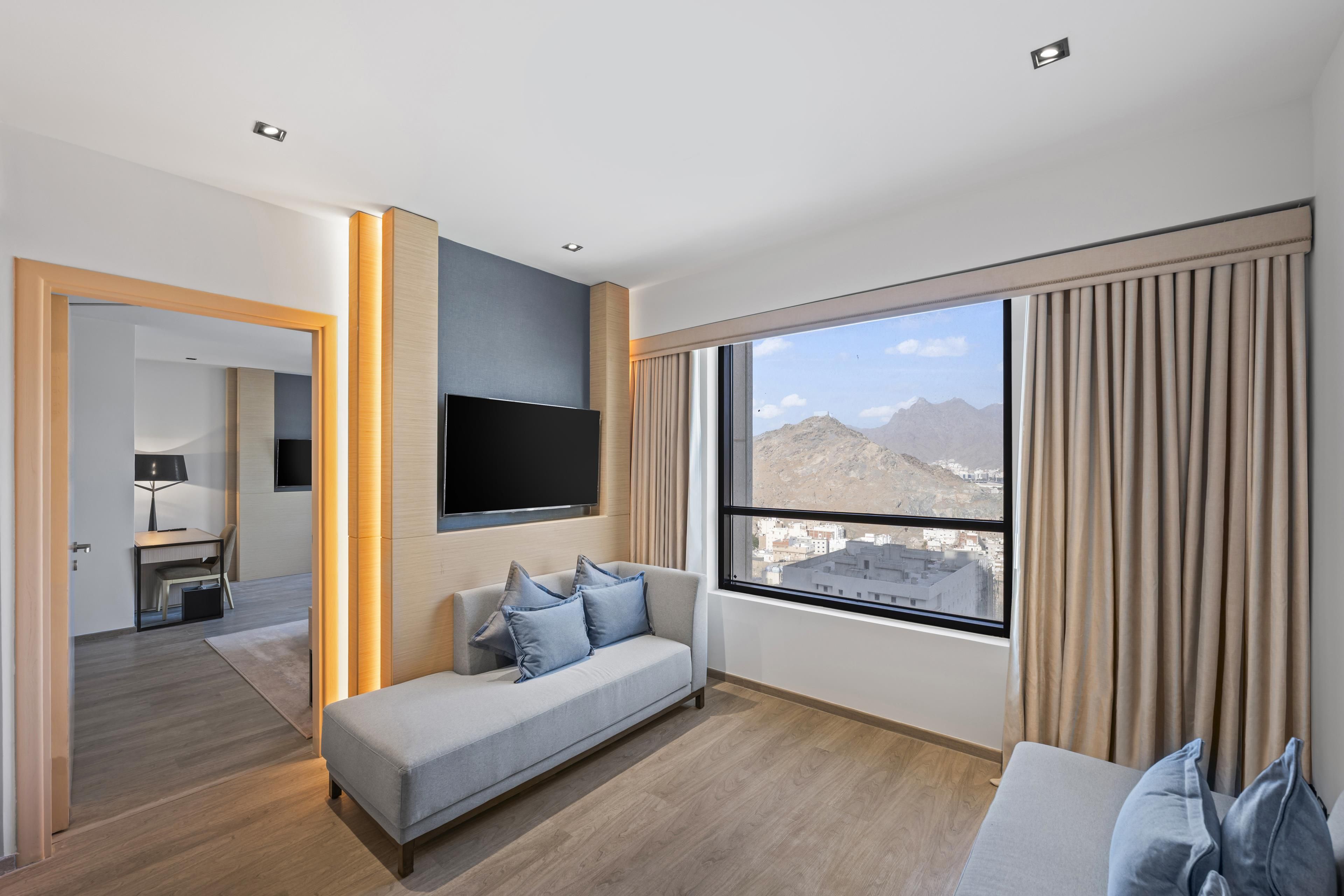A modern hotel room with a sofa, a flat-screen TV mounted on the wall, a large window with curtains, and a view of the cityscape.