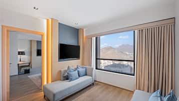 Una habitación de hotel moderna con sofá, televisor de pantalla plana, ventana grande con cortinas y vistas a las montañas.