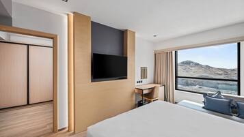 Ein modernes Hotelzimmer mit Bett, Schreibtisch, Fernseher und einem großen Fenster mit Blick auf eine gebirgige Landschaft.