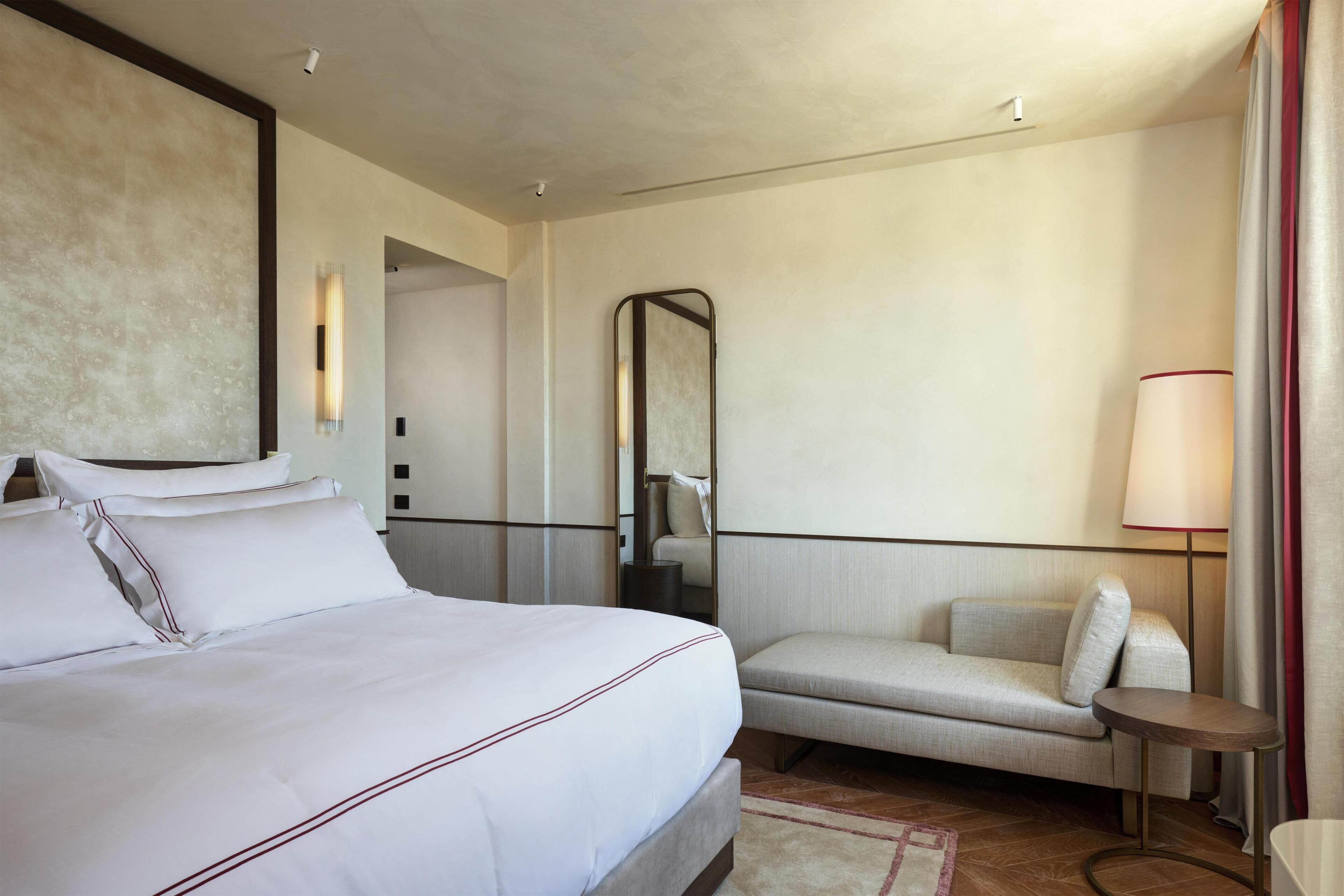 Quarto de hotel com uma cama grande, um sofá pequeno, um espelho, uma mesa de apoio e um abajur.