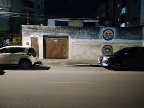 A street at night with two parked cars, a building with a door, and a wall with a circular emblem. - Hospedagem Simples Para Descanso  (Recife)