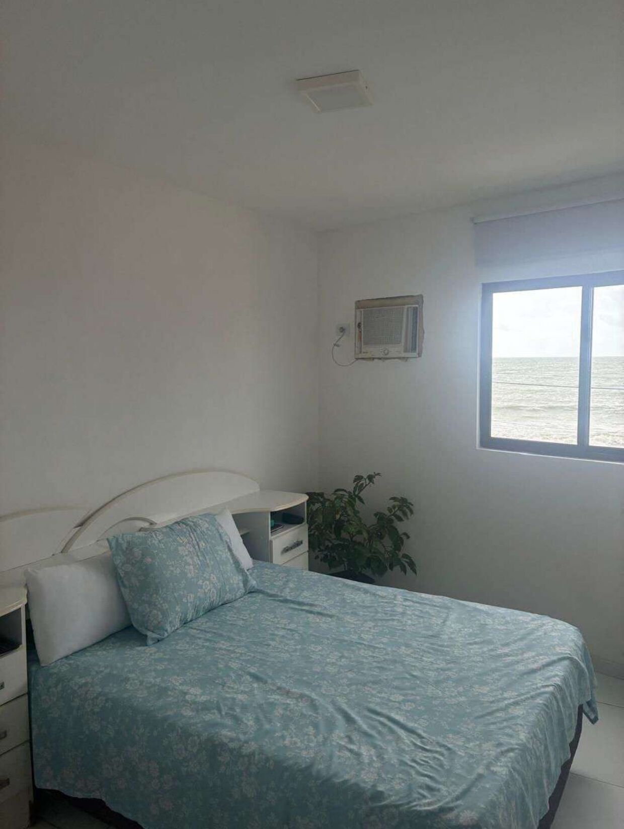Een slaapkamer met een bed, nachtkastje, een klein tafeltje, een kamerplant en een raam met uitzicht op zee.