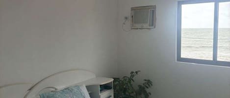 Un dormitorio con cama, mesita de noche, una pequeña mesa, una planta en maceta y una ventana con vista al mar.