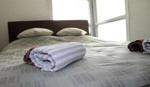 A neatly made bed with a rolled towel and a folded blanket, a window with blinds, and a headboard.