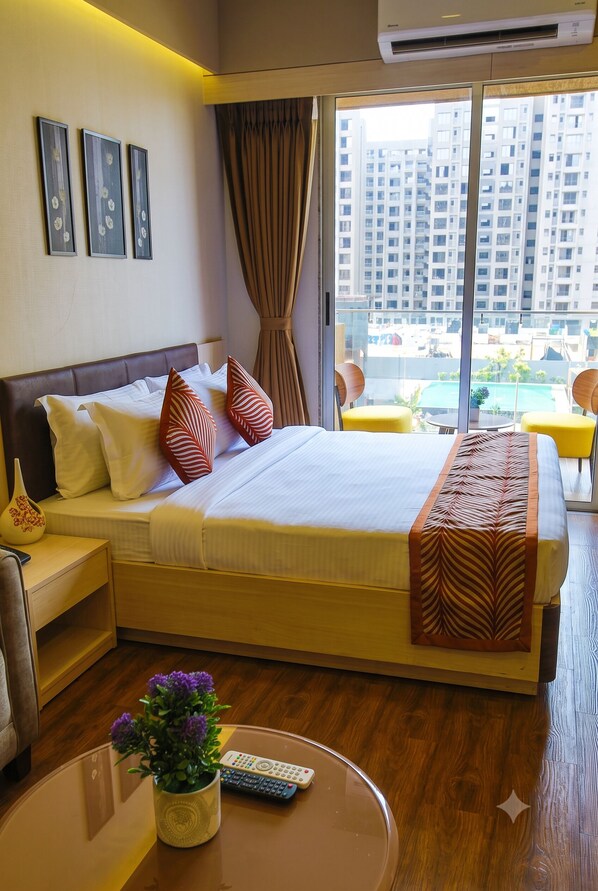 A hotel room with a large bed, a bedside table, a chair, a small table with a vase and remote control, and a window with curtains.