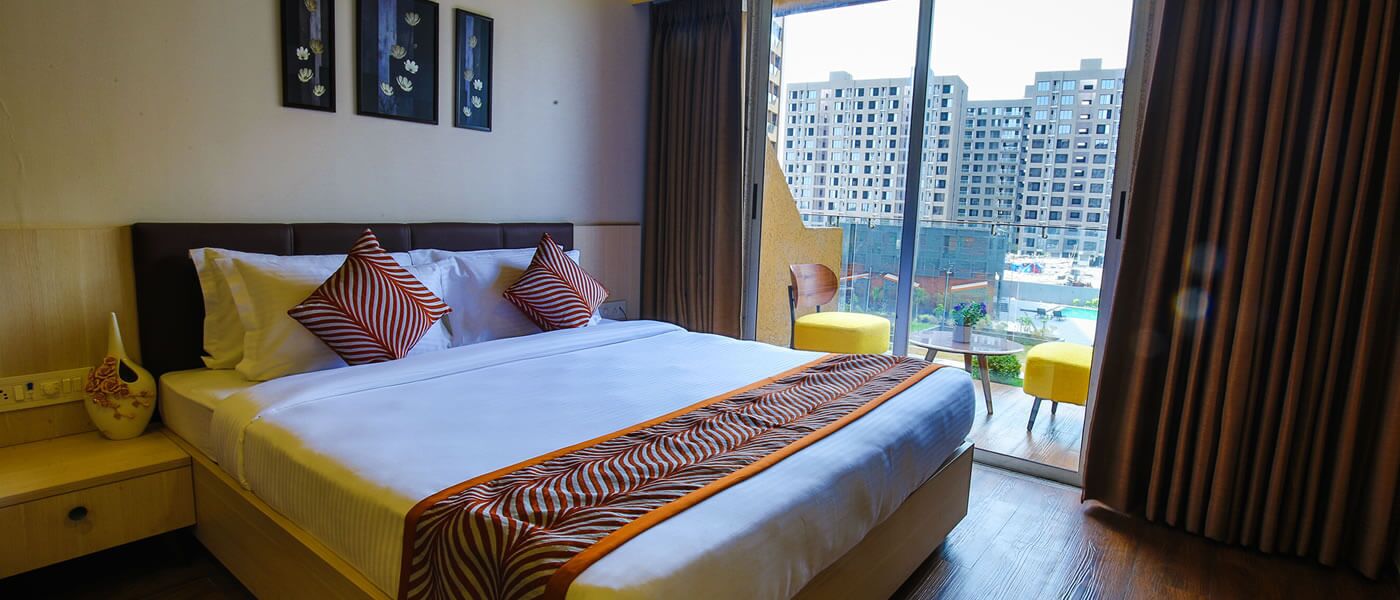 Quarto de hotel com uma cama grande, uma mesa de cabeceira, um vaso e vista para a cidade pela janela.