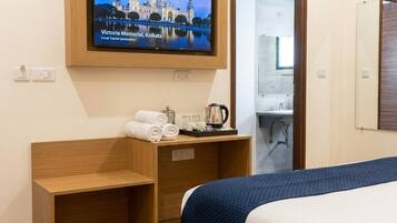 Une chambre d’hôtel avec un lit, un téléviseur fixé au mur, un bureau avec une chaise, une petite table avec des serviettes et une bouilloire, et une salle de bain visible à travers une porte ouverte.