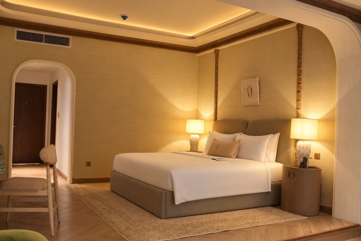 A hotel room with a large bed, two bedside lamps, a wooden headboard, a small bedside table, and a decorative wall piece.