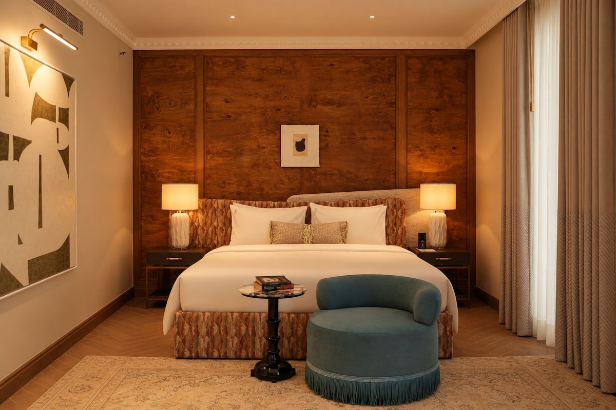 A hotel room with a large bed, two bedside tables, a small round table, and a blue armchair.