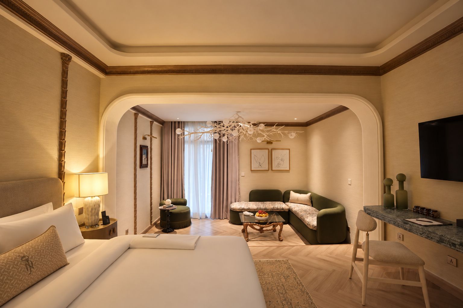 A hotel room with a bed, a sofa, a small table with a fruit bowl, a desk with a chair, and a TV on the wall.