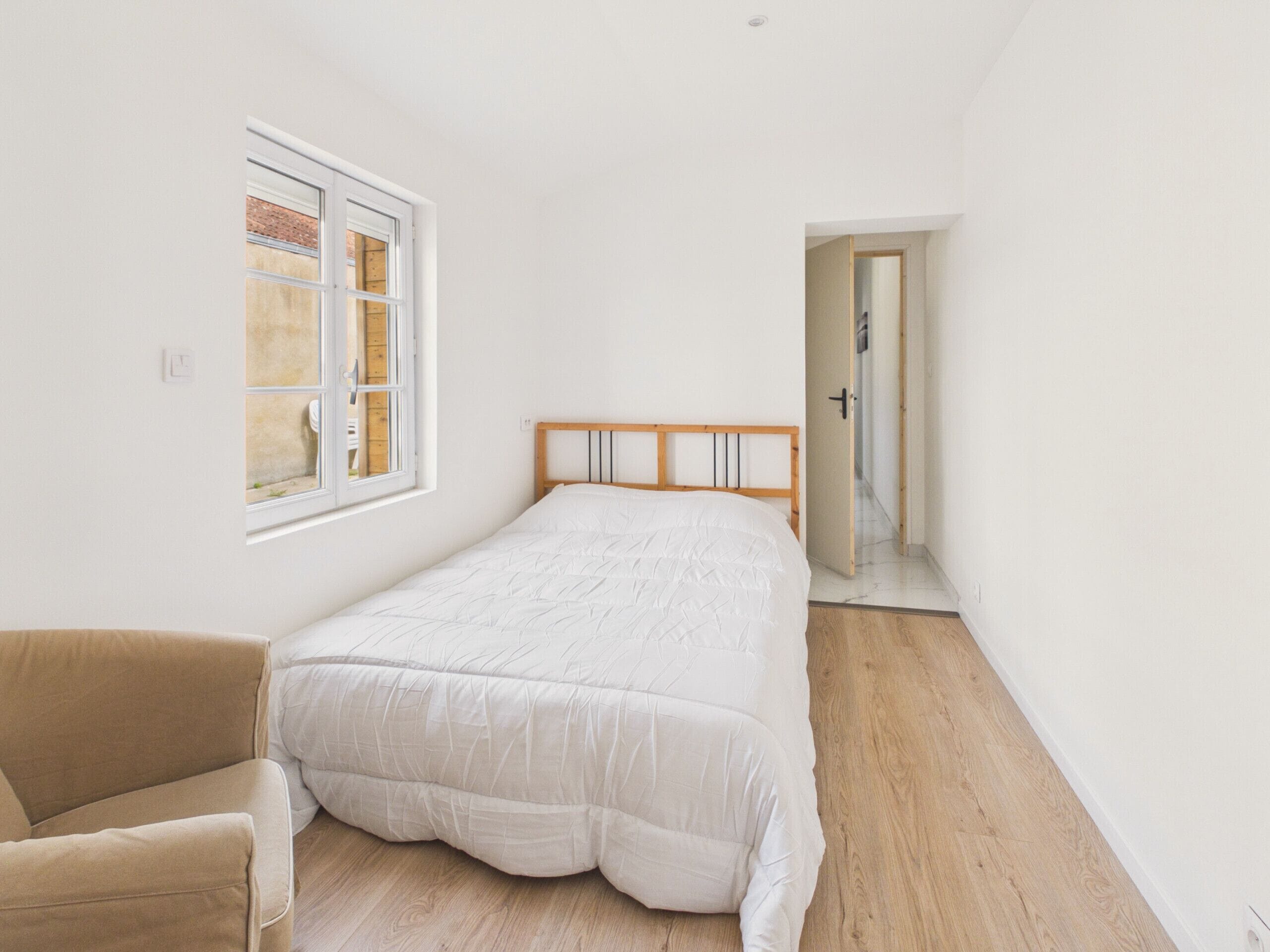 A bedroom with a bed, a chair, a window, and a door leading to another room.
